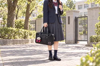 紺ブレザーと膝丈スカートの学生モデルが、さるぼぼ姿のちいかわを1体付けた黒いスクールバッグを持つ全身コーデのAIイメージ